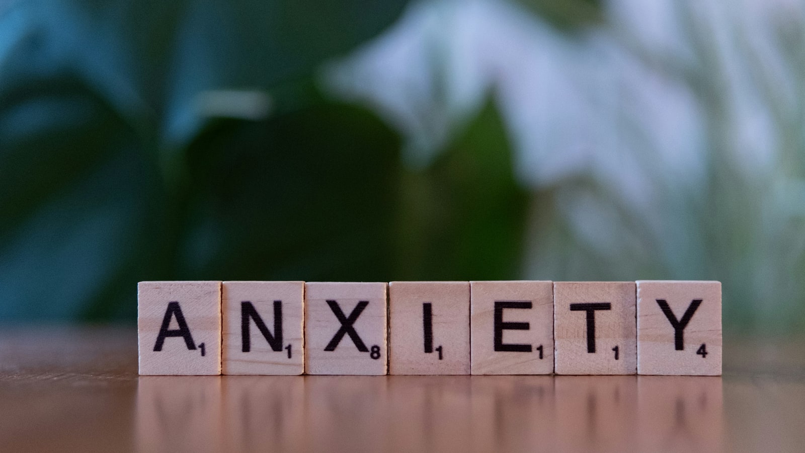 Wooden Scrabble tiles spelling ANXIETY on a wooden surface in front of a softly blurred green plant