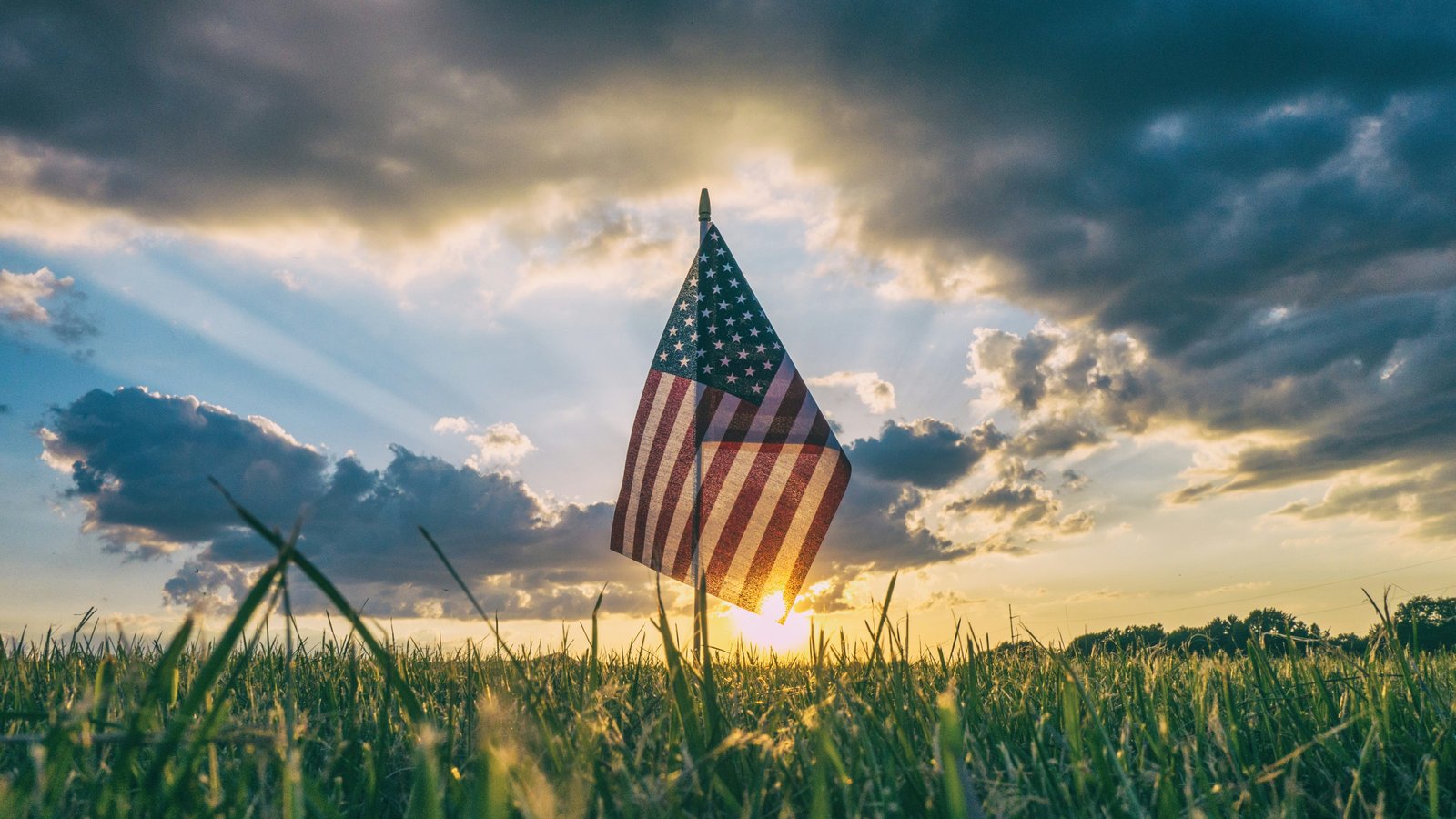 Small American flag planted in a grassy field at sunset with dramatic clouds