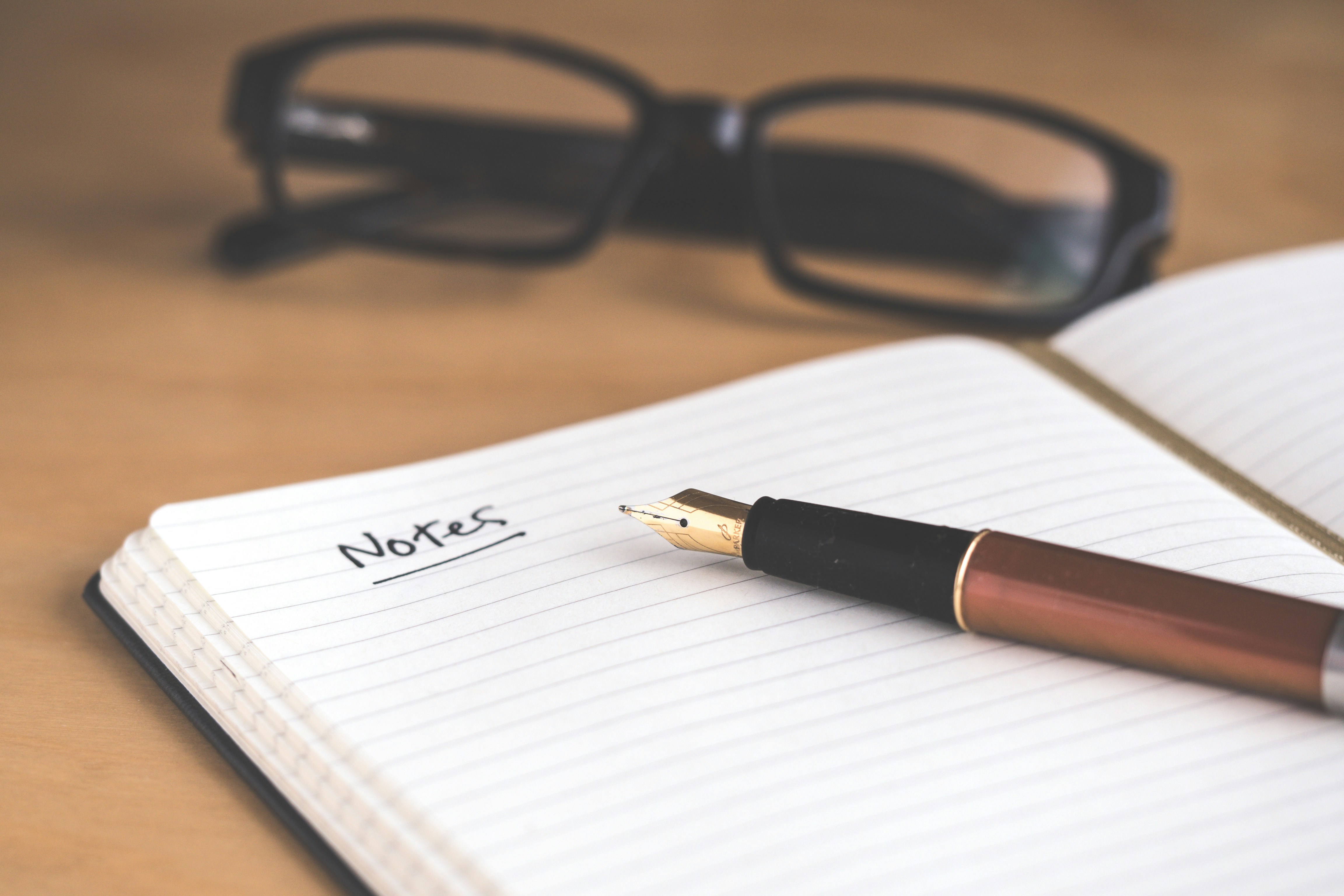 Notebook with 'Notes' written on it, fountain pen, and reading glasses on a wooden desk